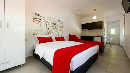 Et hotellrom med to senger har røde sengetepper og puter, et skap til venstre og et kjøkken med mørke skapdører i bakgrunnen. (AI-generert tekst)