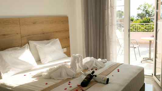 Hotellrom med en dobbeltseng dekorert med håndklær og blomsterblader. En flaske vin og glass ligger på sengen. En balkong er synlig i bakgrunnen. (AI-generert tekst)