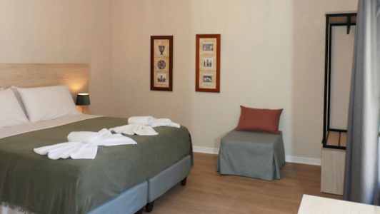 Et hotellrom med en seng, to bilder på veggen, en stol med pute og et åpent garderobeskap. (AI-generert tekst)