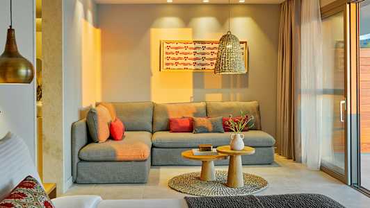 En stue med en grå hjørnesofa, rødlige puter, et par små trebord og et dekorativt teppe. En hengende lampe og et maleri på veggen er også synlig. (AI-generert tekst)