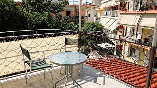 Balkong med glassbord og to metallstoler, med utsikt over grøntområder og omkringliggende bygninger. (AI-generert tekst)