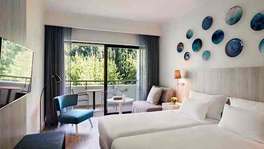 Moderne innredet hotellrom med to senger, en sittegruppe og balkong med utsikt over grøntområder. (AI-generert tekst)