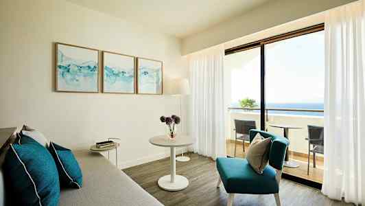 Lyst stue med grå sofa, blå pynteputer og bord, samt utsikt mot balkong og hav. (AI-generert tekst)