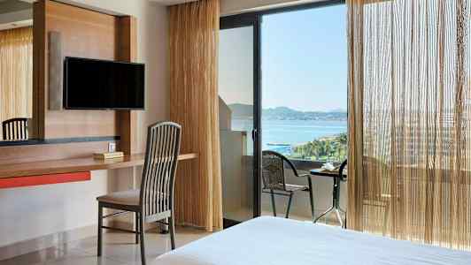 Et hotellrom med skrivebord, stol og TV. Vindu og balkong med havutsikt. (AI-generert tekst)