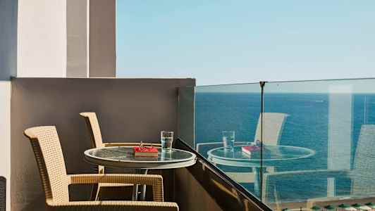Utsikt fra en balkong med to stoler og et bord med et glass vann og en bok foran havet. (AI-generert tekst)