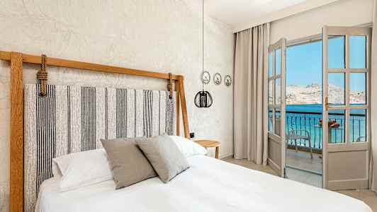 Soverom med en dobbeltseng og en åpning mot en balkong med utsikt over havet. (AI-generert tekst)
