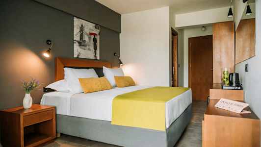 Et soverom med en dobbeltseng med hvite og gule sengetepper, matchende puter, et nattbord med en vase med blomster, og et bilde på veggen over sengen. (AI-generert tekst)