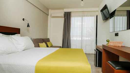 Et hotellrom med en dobbeltseng med hvitt sengetøy og gult sengeteppe, en sofa med puter, et skrivebord med speil og en TV på veggen. Balkongdører med lyse gardiner. (AI-generert tekst)