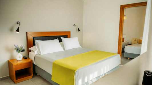 Soverom med en dobbeltseng, to hvite puter og et gult teppe. Et sidebord med en hvit vase og en plante. (AI-generert tekst)