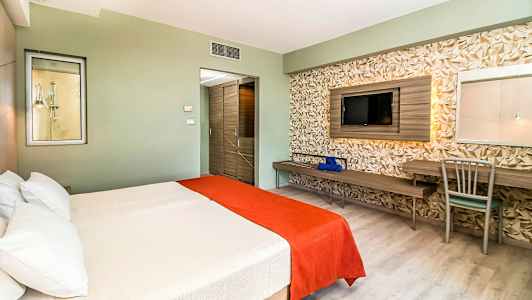 Hotellrom med en dobbeltseng dekket med hvite og oransje sengetepper, en vegg med geometrisk dekor, samt en TV og skrivebord. (AI-generert tekst)