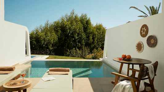 Utendørsområde med to solsenger, et bord med drikke og bok, samt et basseng med trær i bakgrunnen. (AI-generert tekst)