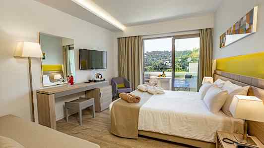 Et hotellrom med seng, skrivebord, TV og en balkong som viser en grønn utsikt. (AI-generert tekst)