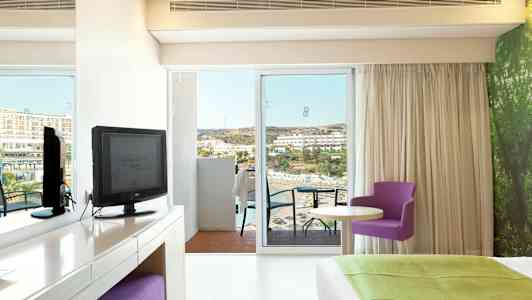 Hotellrom med utsikt mot sjøen, møbler inkludert en TV, et skrivebord, en lilla stol og en stor seng. (AI-generert tekst)