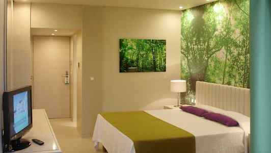 Et hotellrom med en seng, grønn og hvit innredning, en TV og en veggdekorasjon som forestiller en skog. (AI-generert tekst)