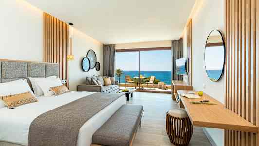 Et stilrent hotellrom med en dobbeltseng, sofa og et skrivebord. Store glassdører åpner seg mot en balkong med utsikt over havet. (AI-generert tekst)