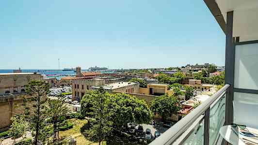 Utsikt fra en balkong over en by med historiske bygninger, trær og en havn med båter. (AI-generert tekst)