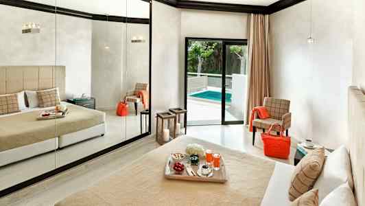 Et hotellrom med en stor speilvegg, en seng med beige sengeteppe og puter, og en balkongdør som fører ut til et basseng. (AI-generert tekst)