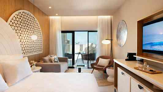 Et moderne hotellrom med lyse møbler og dekorasjoner, inkludert en seng, sofa, stol og TV. Rommet har store glassdører som fører ut til en balkong. (AI-generert tekst)