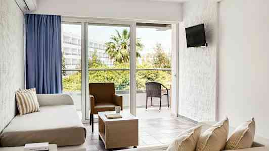 Et lyst hotellrom med sittegruppe, sofaer, en stol, et salongbord og store vinduer som vender ut mot en balkong med utemøbler i et frodig miljø. (AI-generert tekst)