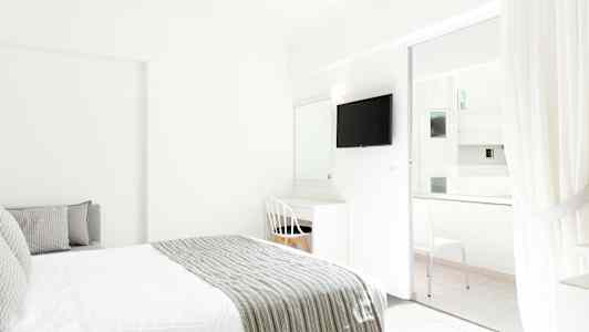 Interiør av et lyst hotellrom med en seng og skrivebord ved siden av en hvit stol. En TV er montert på veggen, og glassdører fører til en balkong. (AI-generert tekst)