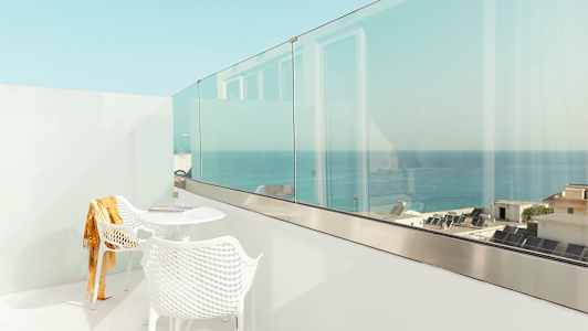 En hvit balkong med to stoler og et bord, med utsikt over havet og bygninger. (AI-generert tekst)