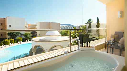 Bilde av en balkong med et boblebad og sittemøbler. Utsikt over et basseng og omkringliggende bygninger. (AI-generert tekst)