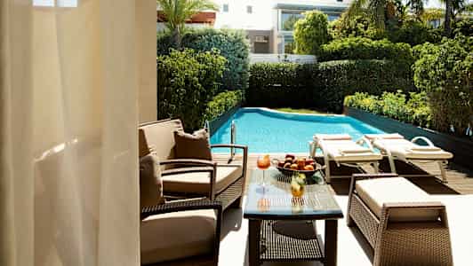 Utsikt over et basseng med solsenger og hage, samt en sittegruppe med bord. (AI-generert tekst)