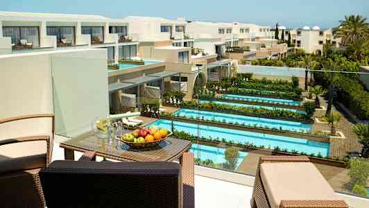 Hotellområde med flere bassenger og grøntområder, samt en terrasse med møbler og frukt. Utsikt over en moderne bygning. (AI-generert tekst)