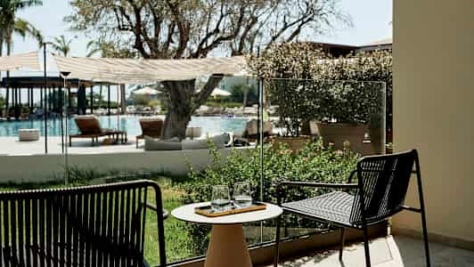 Utsikt mot et utendørs basseng med stoler og planter i forgrunnen. (AI-generert tekst)