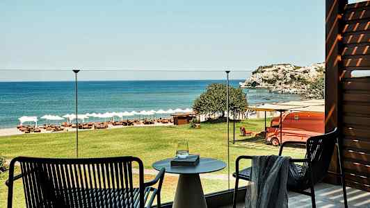 Utsikt fra en terrasse over grønt gress, en strand med solsenger og parasoller samt havet og klipper i bakgrunnen. (AI-generert tekst)