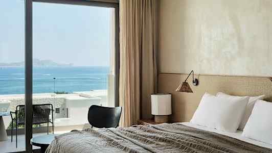 Et hotellrom med seng, bord og stol, med havutsikt gjennom store vinduer. (AI-generert tekst)