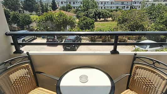 En balkong med to stoler og et rundt bord med en askebeger, med utsikt over en parkeringsplass og grønne omgivelser. (AI-generert tekst)