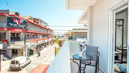 En balkong med bord, stol og blomster i forgrunnen, samt utsikt over en gate med butikker og bygninger. (AI-generert tekst)