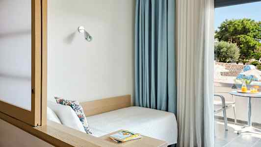 Et lite rom med en seng ved et vindu og utsikt over en uteplass med bord og stoler. (AI-generert tekst)