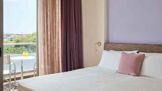 Et hotellrom med en oppredd seng, lilla vegger og gardiner, samt en balkong med bord og to stoler med utsikt mot vegetasjon. (AI-generert tekst)