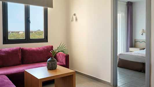 Et rom med lilla hjørnesofa, et bord med en vase og et tilstøtende soverom synlig gjennom en døråpning. (AI-generert tekst)