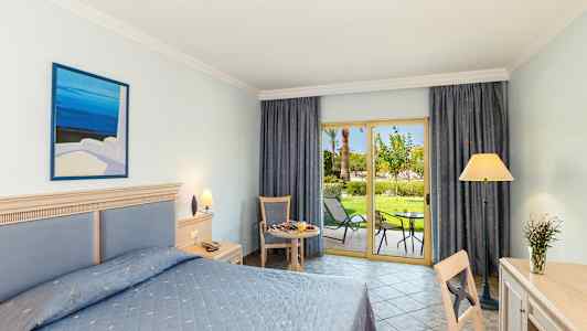 Hotellrom med dobbeltseng, skrivebord, sittemøbler og åpen balkongdør mot en hage. (AI-generert tekst)