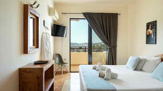 Et hotellrom med en seng, TV, stol og utsikt over en balkong. (AI-generert tekst)