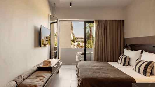 Et hotellrom med en seng, en benk med en hatt og utsikt mot en balkong med trær. (AI-generert tekst)