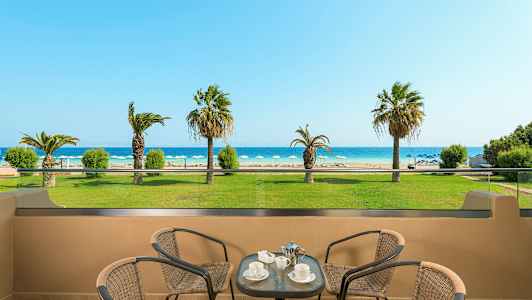 Utsikt fra en terrasse mot en strand med solsenger og palmer, med bord og stoler i forgrunnen. (AI-generert tekst)