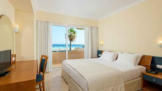 Hotellrom med en dobbeltseng, skrivebord, stol og utsikt over en strand gjennom en glassdør. (AI-generert tekst)