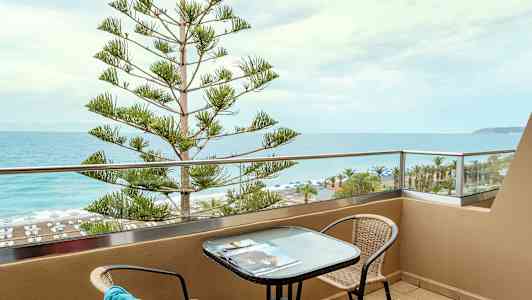 En balkong med utsikt over havet og stranden, med et bord og stoler. (AI-generert tekst)