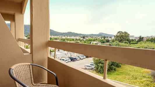En balkong med en stol og utsikt over en parkeringsplass samt grøntområder og fjell i det fjerne. (AI-generert tekst)