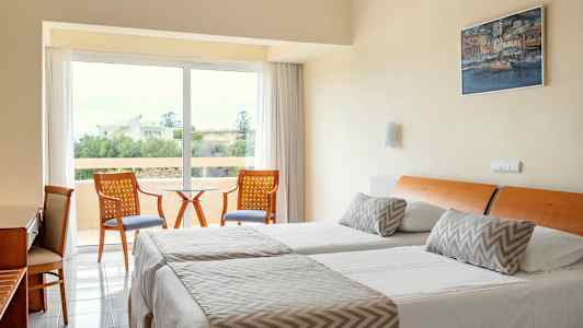 Et hotellrom med to enkeltsenger, en sittegruppe og utsikt til grønne omgivelser. (AI-generert tekst)