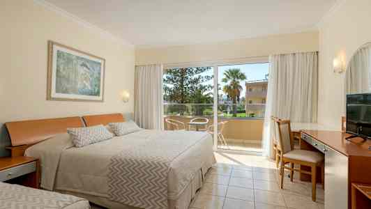 Et hotellrom med oppredd seng, skrivebord og stoler ved siden av en glassdør som fører til en balkong. (AI-generert tekst)