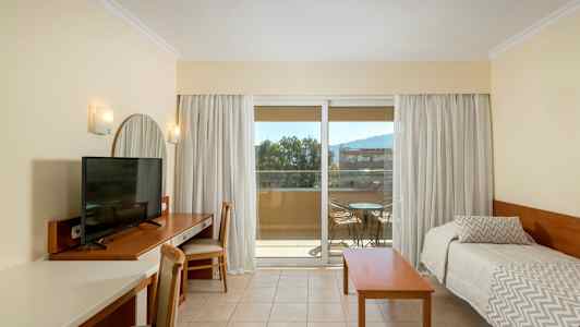 Et hotellrom med en TV på en skrivebord, to enkeltsenger og en balkong med utemøbler. (AI-generert tekst)