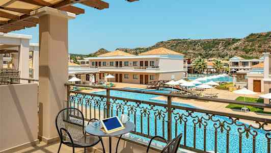 Utsikt fra en balkong mot et basseng og hotellbygninger med solsenger og parasoller. (AI-generert tekst)