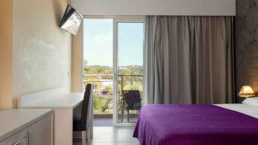 Et hotellrom med et skrivebord, en stol, en TV og en seng med lilla sengeteppe. Rommet har en balkong med utsikt. (AI-generert tekst)