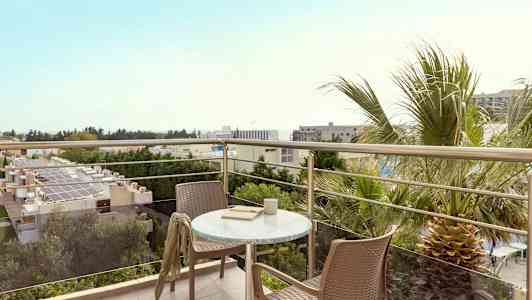 Utsikt fra en balkong med bord og to stoler, omgitt av trær og bygninger. (AI-generert tekst)