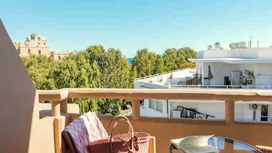 Utsikt fra en balkong mot trær og bygninger. Et bord med en bok og en stol med en veske i forgrunnen. (AI-generert tekst)